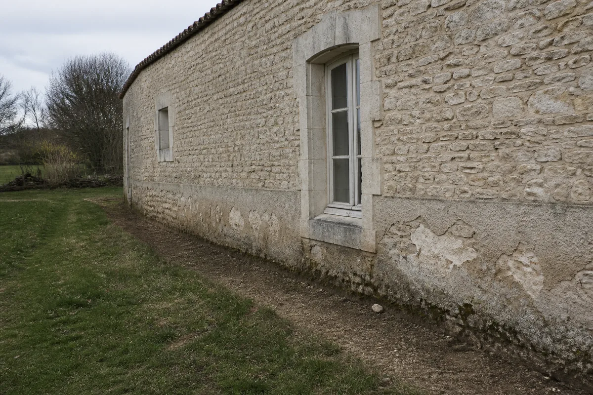 Mur ancien en moellons calcaires avec enduit à la chaux et sol décaissé le long de la façade